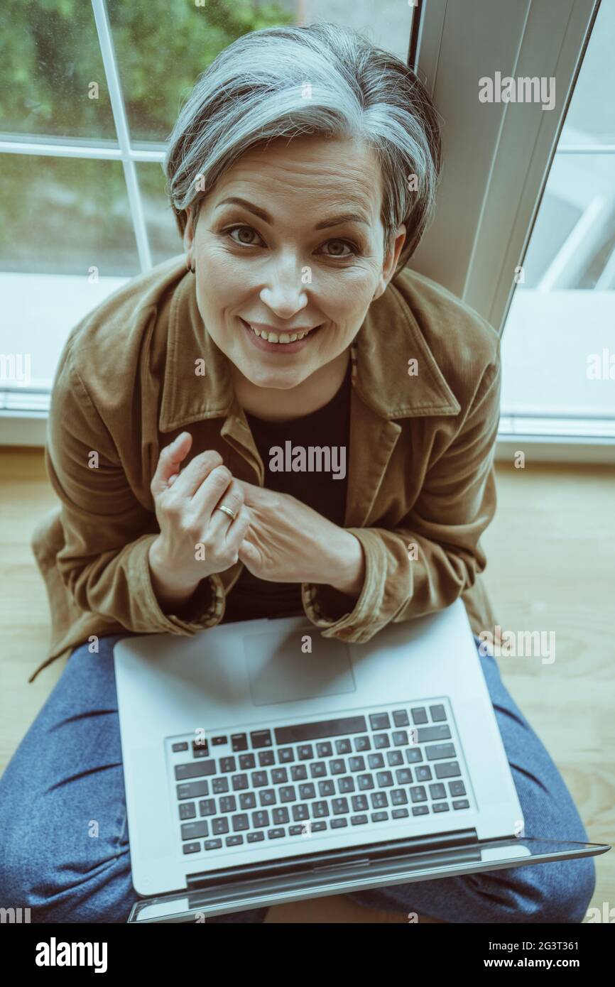 Charming mature woman looks up at camera folding hands while holding laptop on her knees. Aged businesswoman works sitting at si Stock Photo