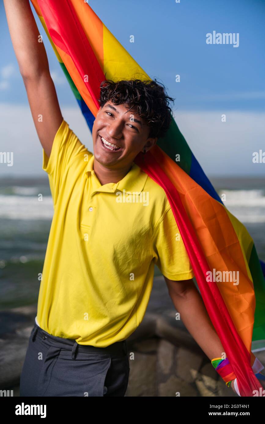 Young gay man proudly holding the lgbt flag in his hands in front of the sea, Inclusive Stock Photo