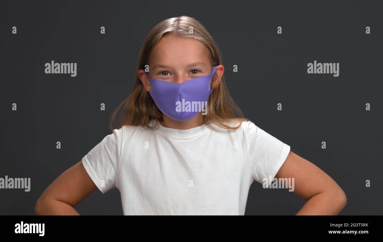 Little blonde girl in lilac mask smiles against gray background. Caucasian child looking at camera winks. Studio shot Stock Photo