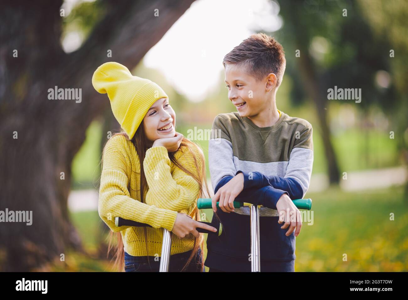 Twins 10 years old in bright clothes spend time actively together in autumn park riding kick scooters. Happy children. Eco trans Stock Photo