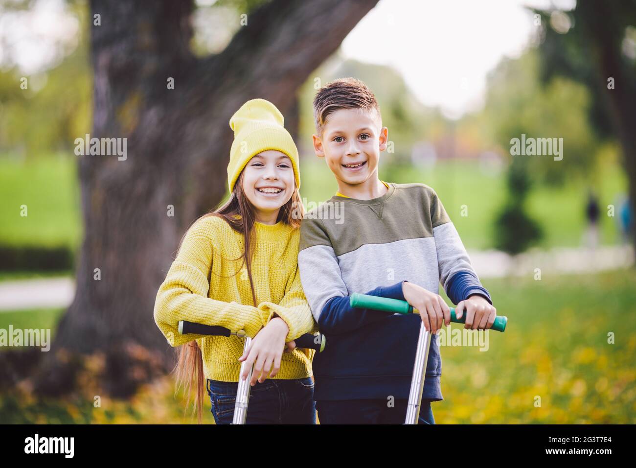 Caucasian children twins boy and girl schoolchildren age pose together in sunny weather in autumn park after riding on kick scoo Stock Photo
