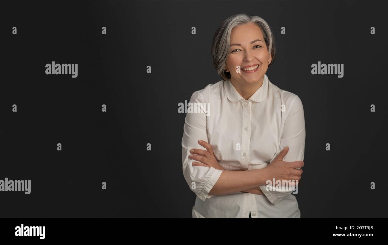 Senior woman laughing on grey background. Portrait of mature grey haired woman in white casuals looking happy on camera Stock Photo