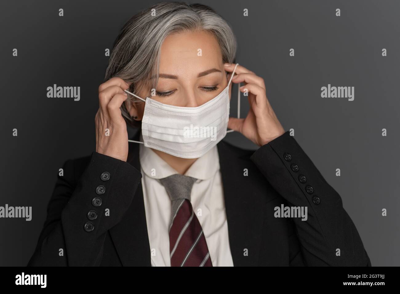 Grey-haired middle-aged business woman putting on a protective mask with eyes closed isolated on grey background. Portrait of mo Stock Photo