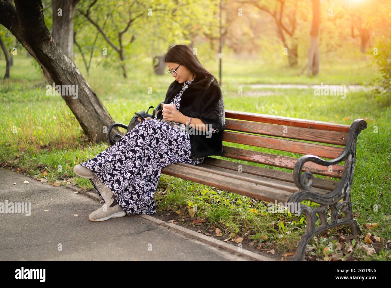 Woman of Asian ethnicity in beautiful long dress sits at park on old rusty bench and reads book using her gadget or checks socia Stock Photo