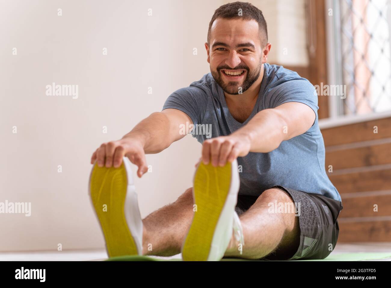 Young happy smiling fitness man doing stretching exercises at home. Amateur sports man in workout activity sitting on pad and st Stock Photo