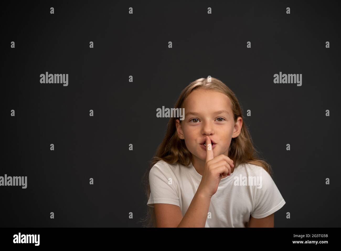 Keep quiet 8,10 years old girl holding her finger on her mouth telling everyone to keep silence wearing white t shirt smiling at Stock Photo