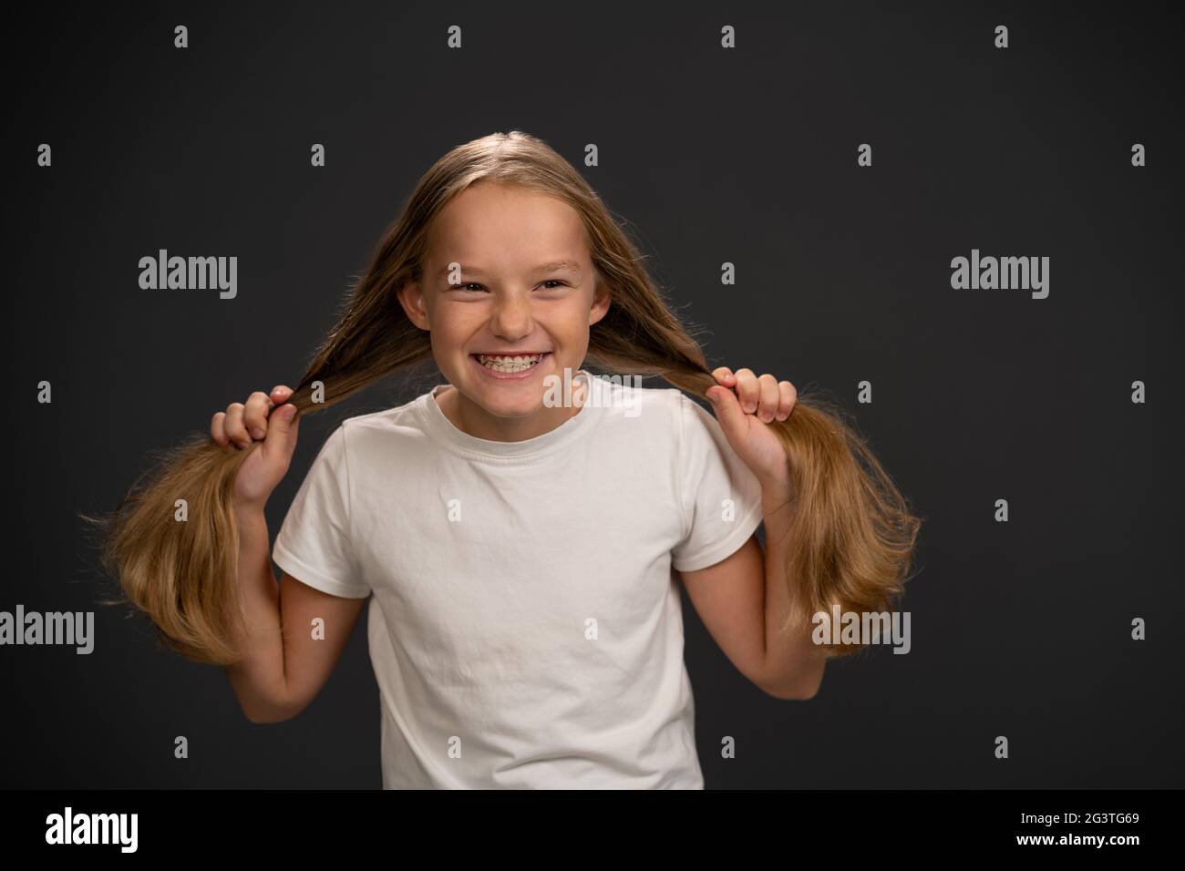 Happy smiling and holding two pony tails 8,10 years old girl holding her hair wearing white t shirt smiling a little grinning at Stock Photo