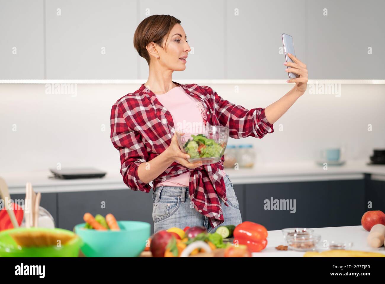 Pretty housewife taking selfie or making a video call using her smartphone while cooking fresh salad wearing a plaid shirt with Stock Photo