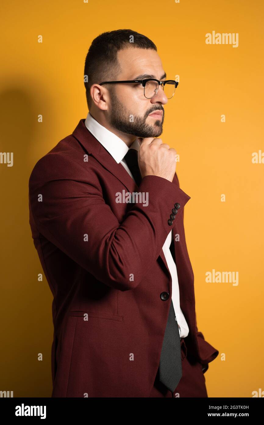 Handsome young man eye glasses wearing burgundy suit looking seriously away with hand folded touching chin standing sideways iso Stock Photo