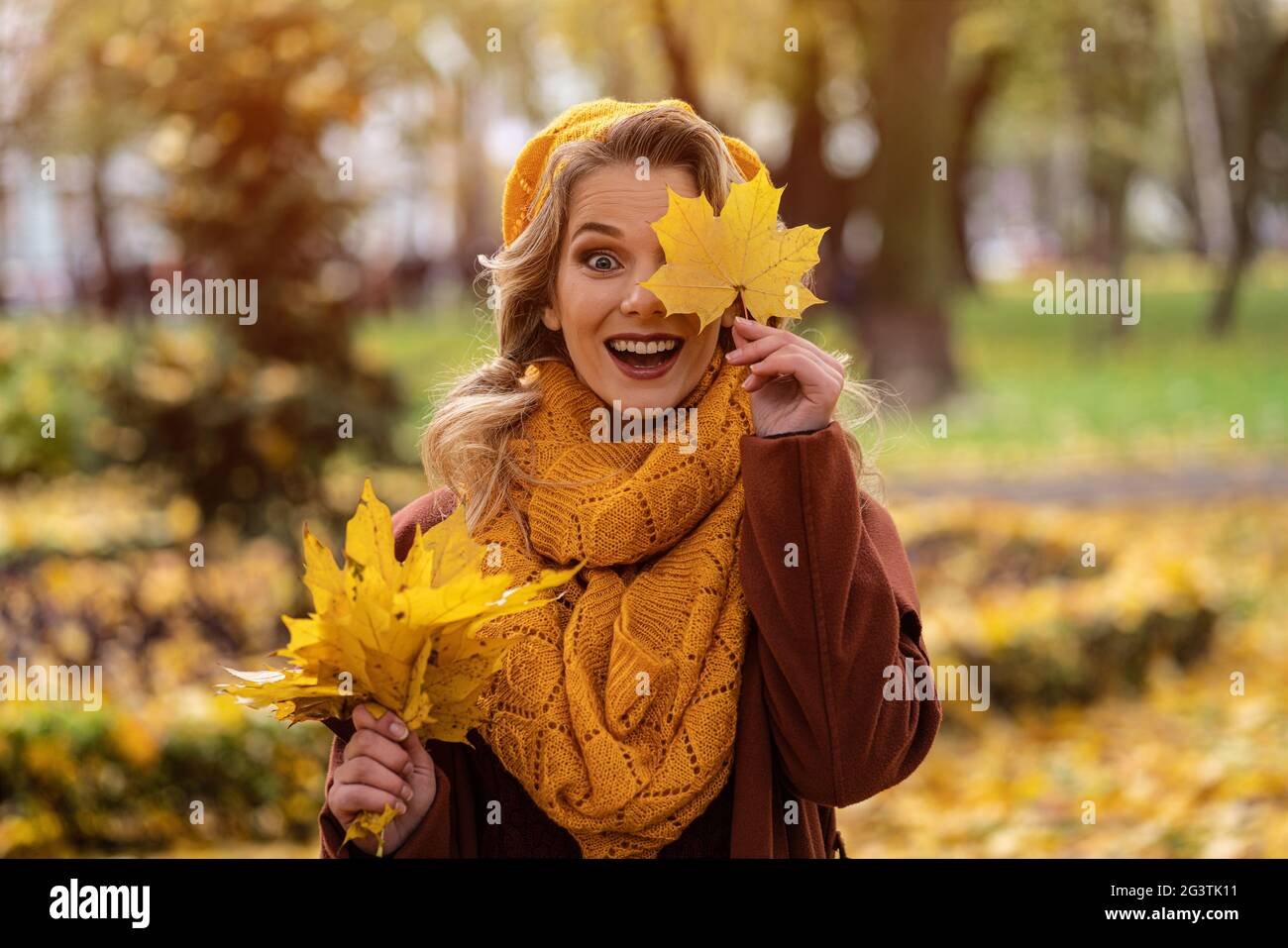Cheerful young woman looking thru yellow fallen leaf in knitted beret with autumn leaves in hand and fall yellow garden or park. Stock Photo