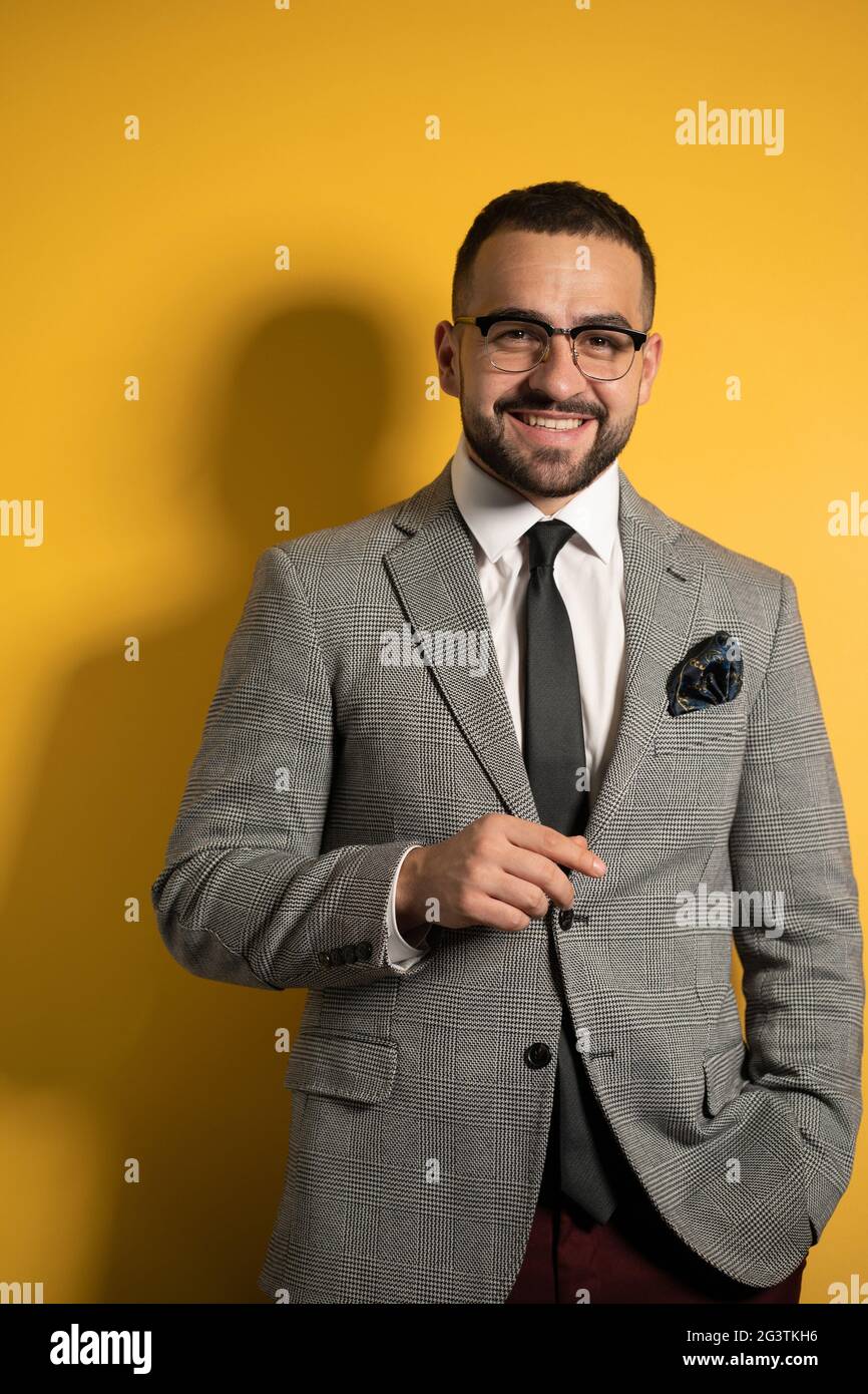 Handsome bearded smiling elegant man wearing eye glasses in formal wear with one hand lifted standing isolated on yellow backgro Stock Photo