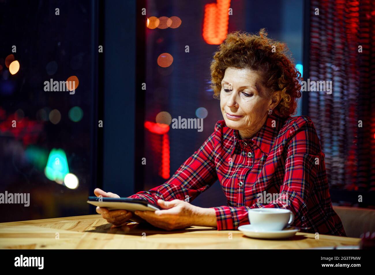 Mature woman with blond curly hair checks mail and social media notifications via tablet, smiling female holding tablet, watchin Stock Photo