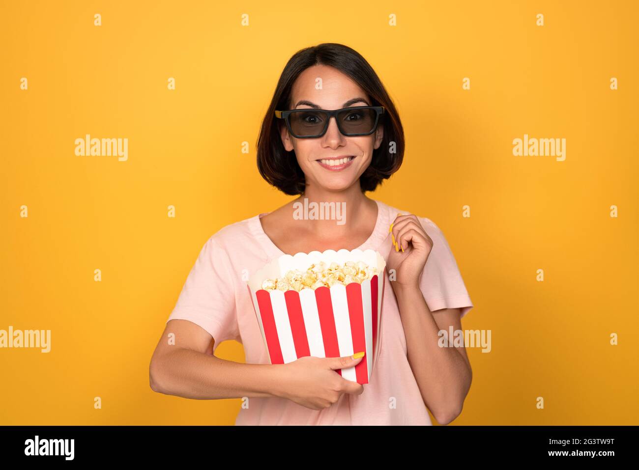 Bucket of popcorn and stereo 3D glasses in hands of a young brunette woman. Cinema concept. Isolated on yellow background Stock Photo
