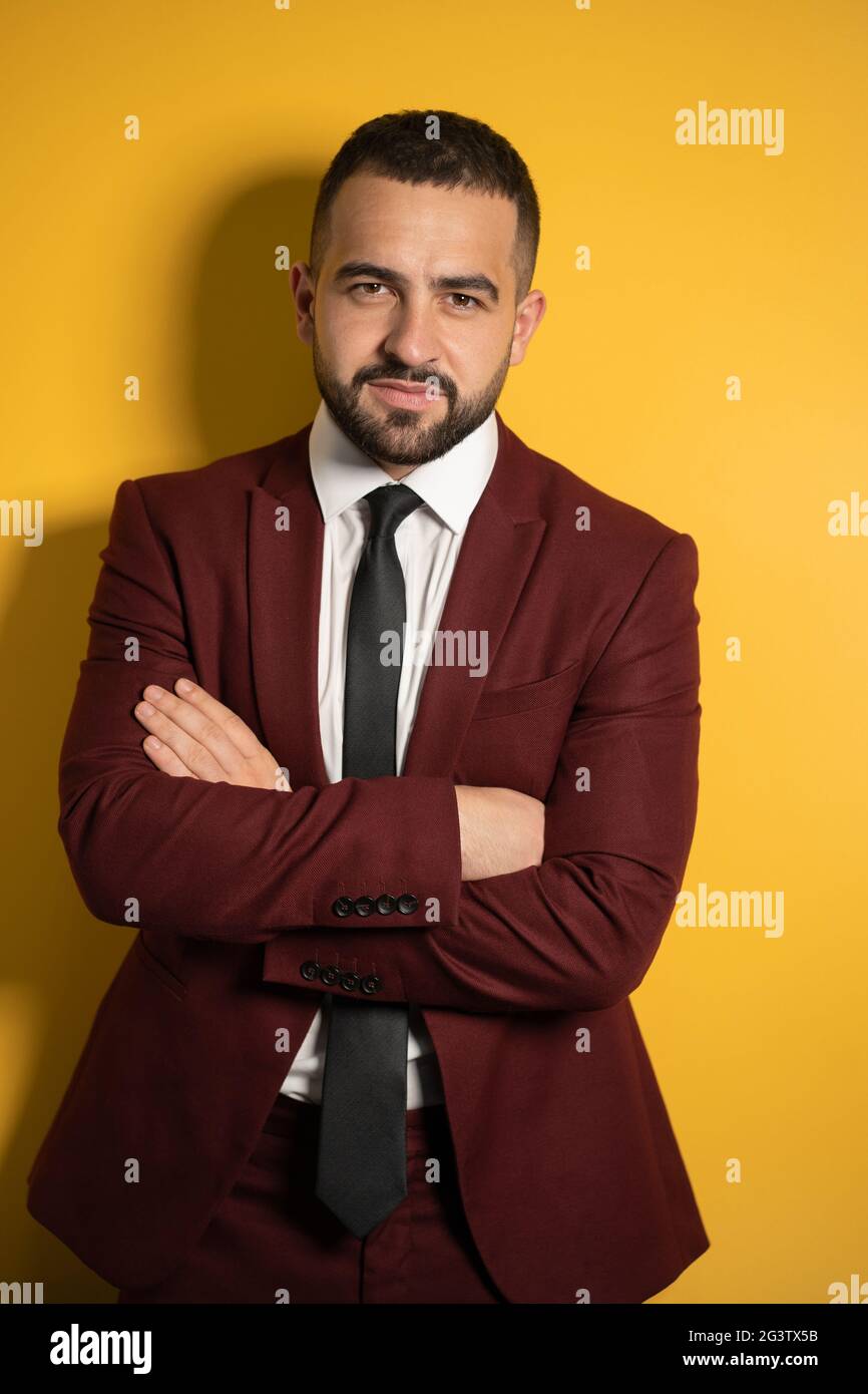 Half-length portrait of handsome man in burgundy color suit looking at camera with hands folded isolated on yellow background. V Stock Photo