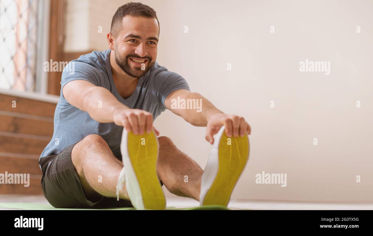 Happy smiling athletic young man stretching out training at home. Amateur sports man in workout activity sitting on pad and stre Stock Photo