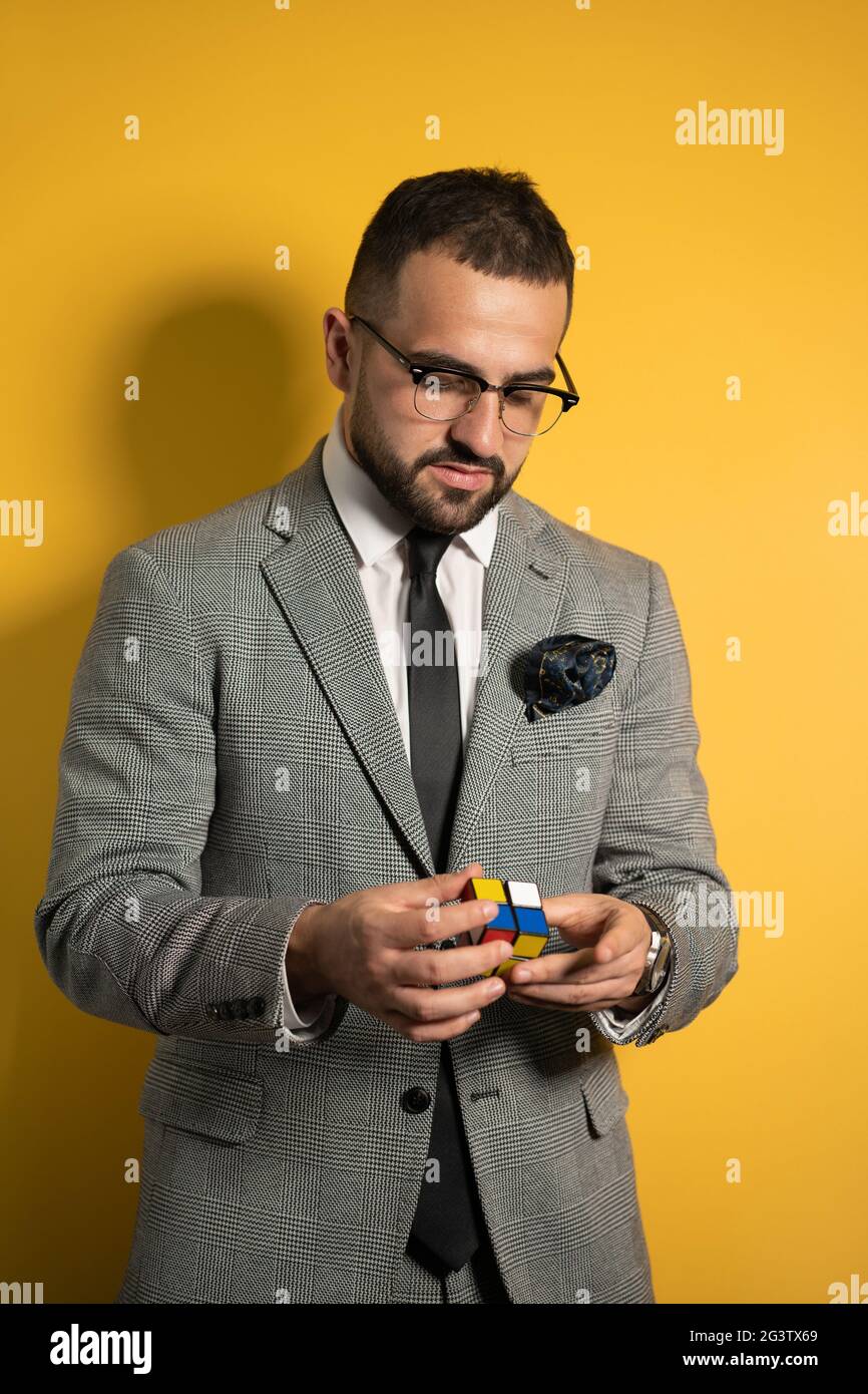 Young handsome man in eye glasses wearing a suite solving a Pocket Cube 2x2x2 rotatable puzzle in hands while looking down on it Stock Photo