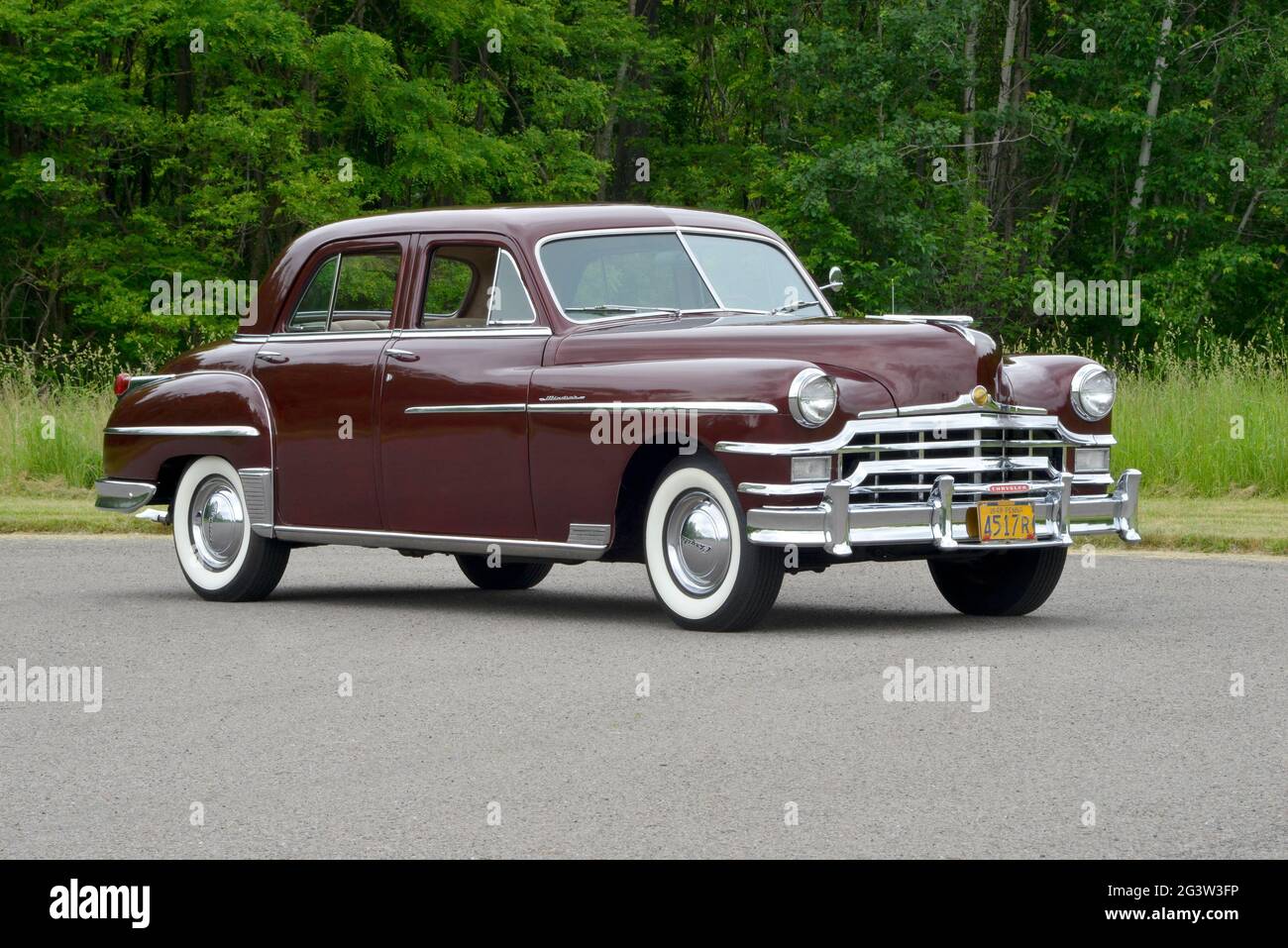 A three-quarter-front view of a 1949 Chrysler Windsor four-door sedan before a wooded background in soft light. Stock Photo