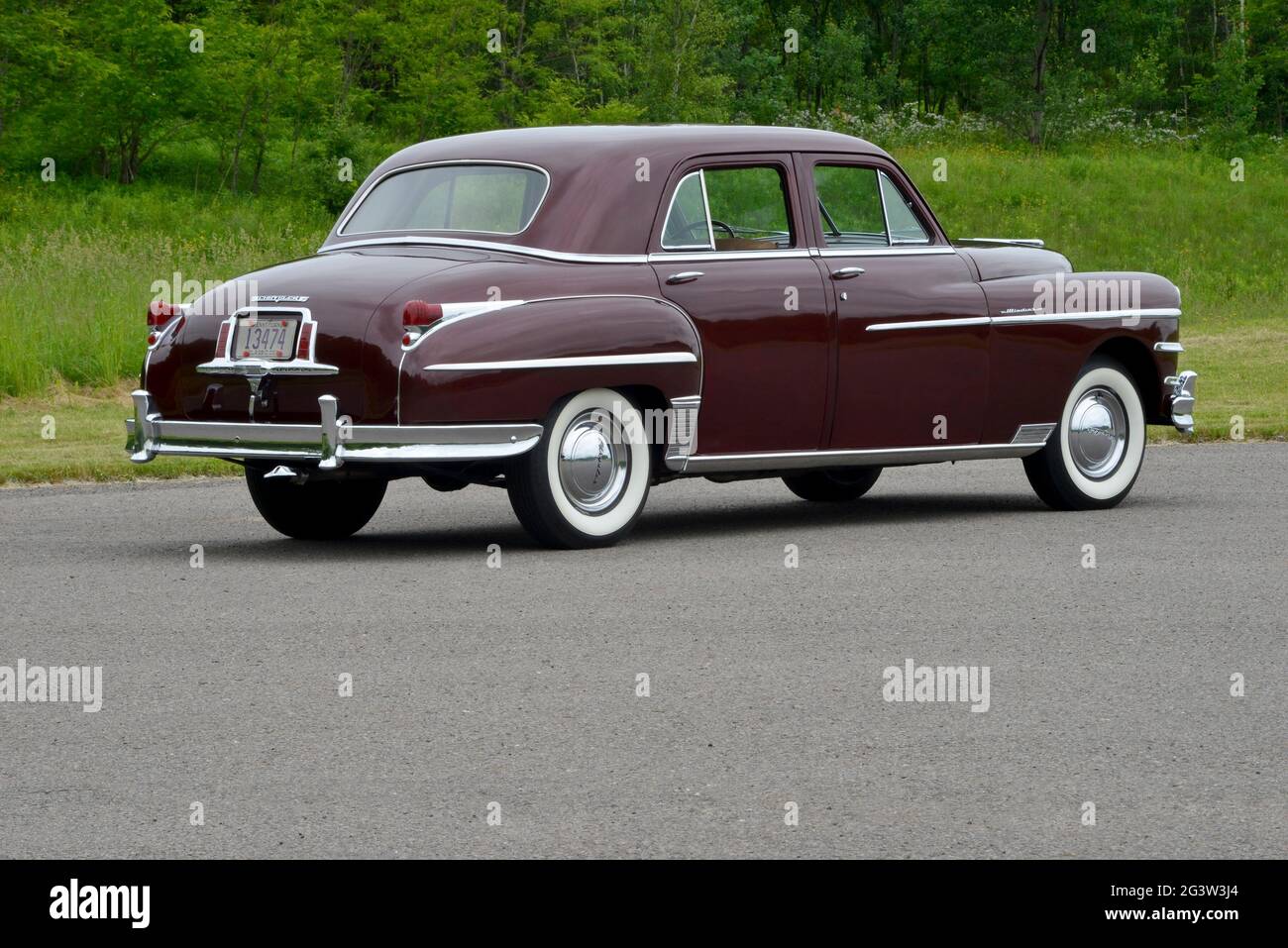 A three-quarter-rear view of a 1949 Chrysler Windsor four-door sedan before a wooded background in soft light. Stock Photo