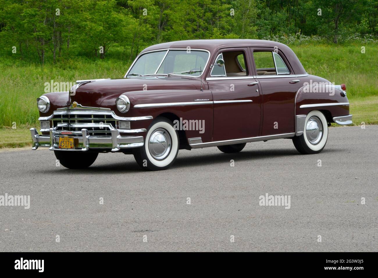 A three-quarter-front view of a 1949 Chrysler Windsor four-door sedan before a wooded background in soft light. Stock Photo