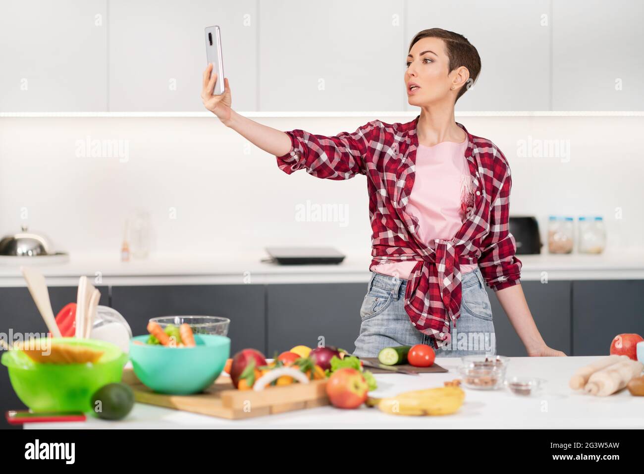 Pretty woman taking selfie or making a video call using her smartphone holding it in outstretched arm while cooking fresh salad Stock Photo
