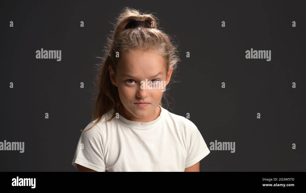 Wicked or malign little girl with pony tail hair looking at the camera wearing white t-shirt and black pants isolated on black b Stock Photo