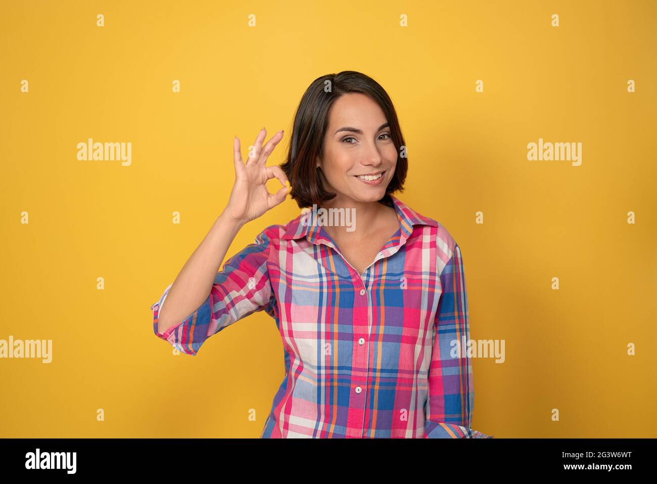 Happy girl showing OK gesture with one raised hand smile beautiful woman dressed in a plaid shirt and dark hair on yellow backgr Stock Photo