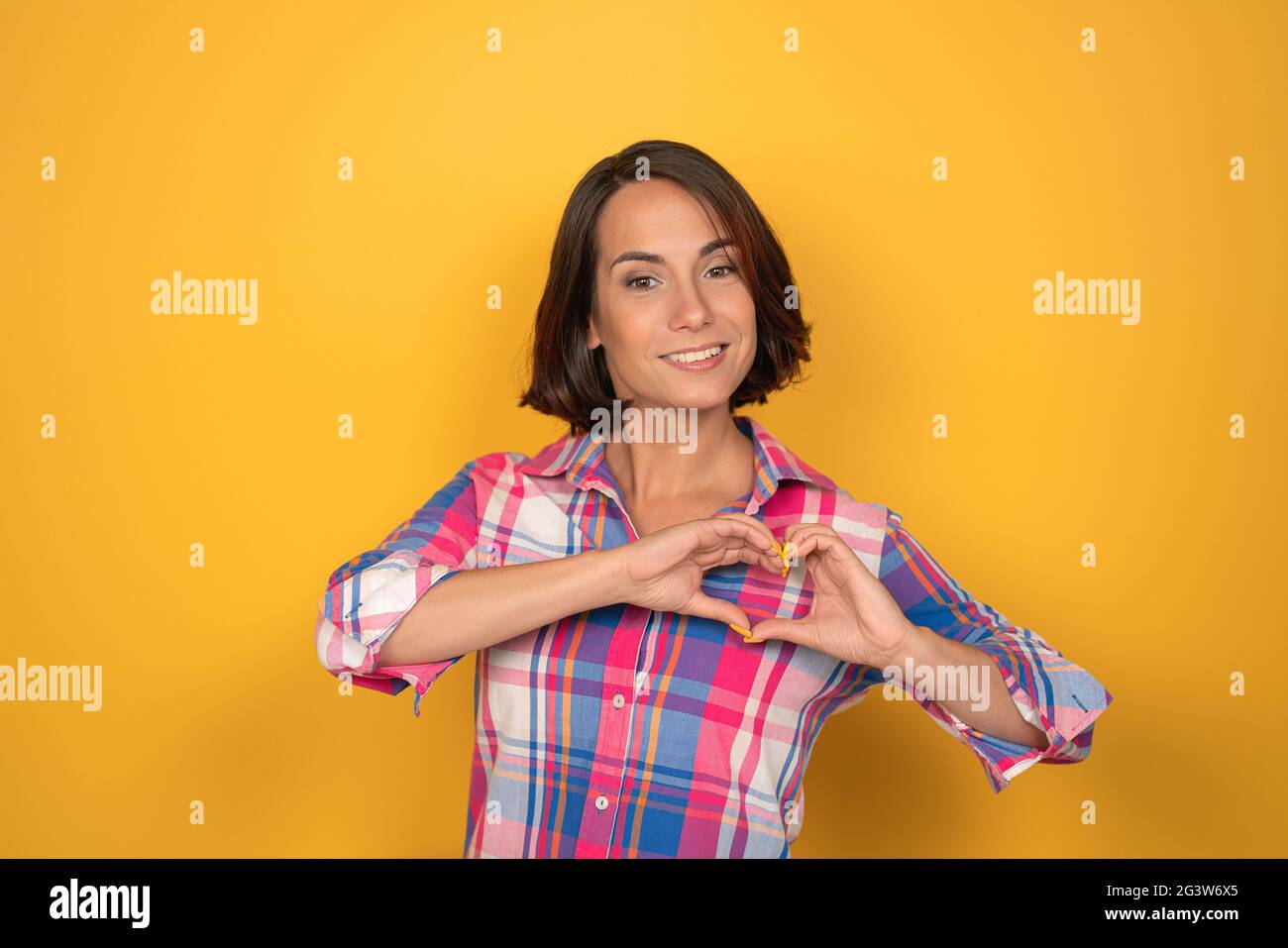 OK or Okay sign with one raised hand smile beautiful woman dressed in a plaid shirt and dark hair on yellow background. Human em Stock Photo