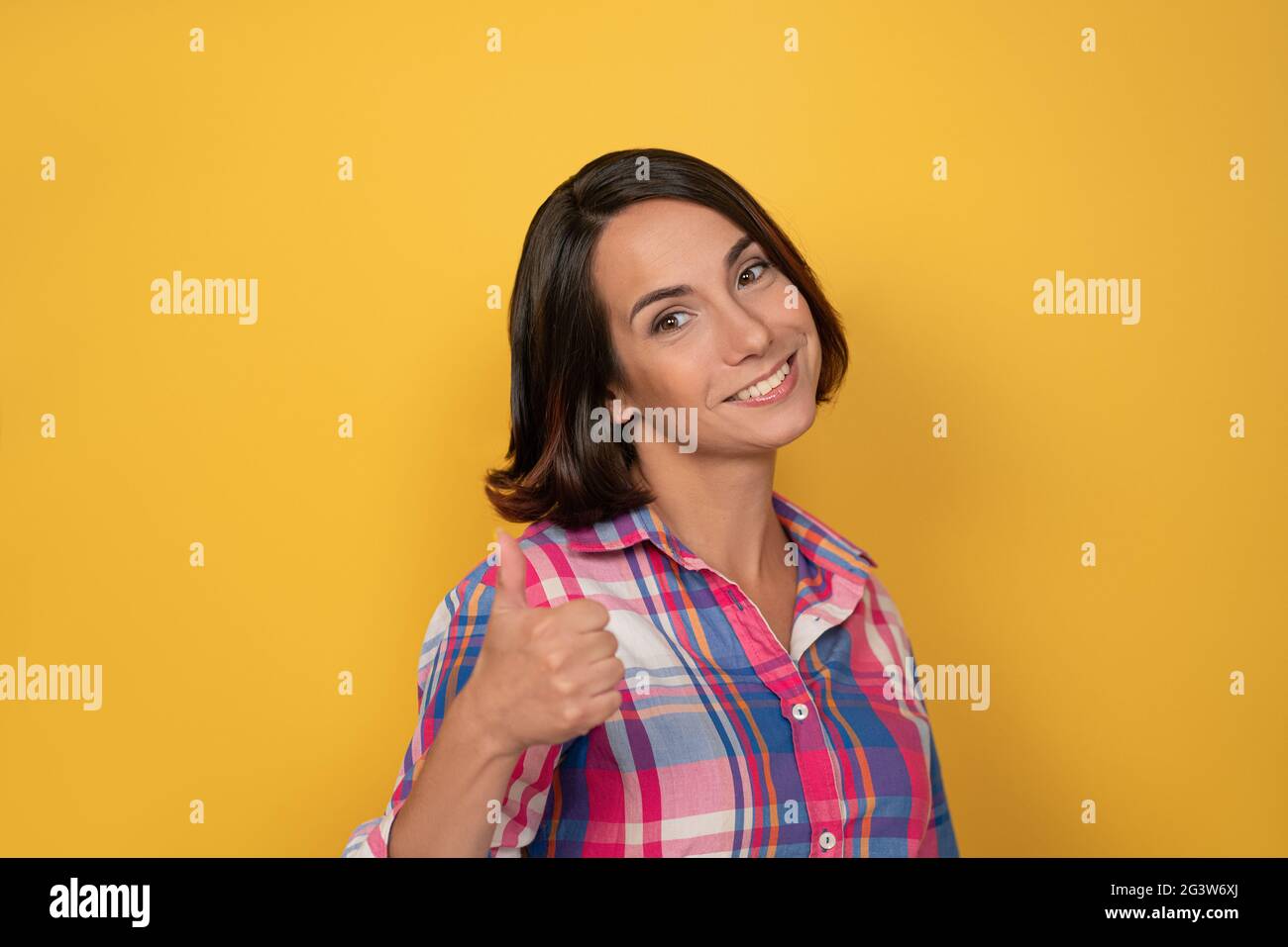 Raising thumb up gesture with hand smile beautiful woman dressed in a plaid shirt and dark hair on yellow background. Human emot Stock Photo