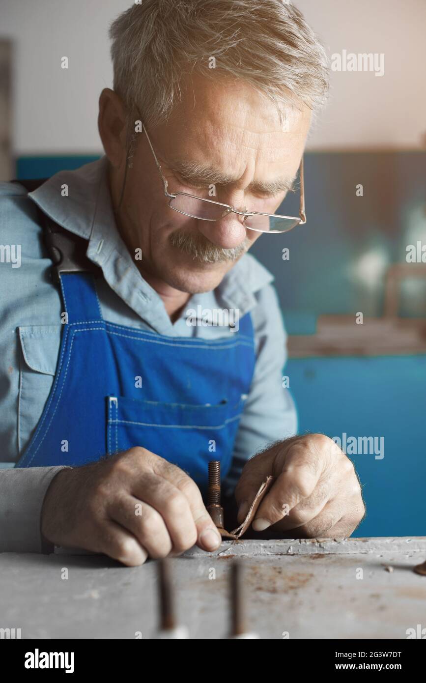 An elderly master with glasses at work. A white old man of Caucasian appearance sits at a table and works with his hands Stock Photo