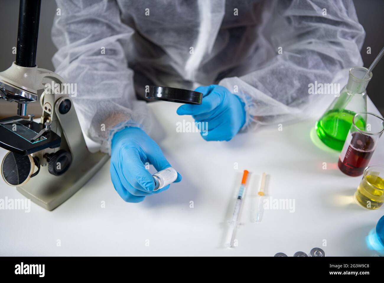 Scientist in hazmat suit and goggles holding vaccine bottle and magnifying glass, looking and research. Epidemiology virus test. Stock Photo