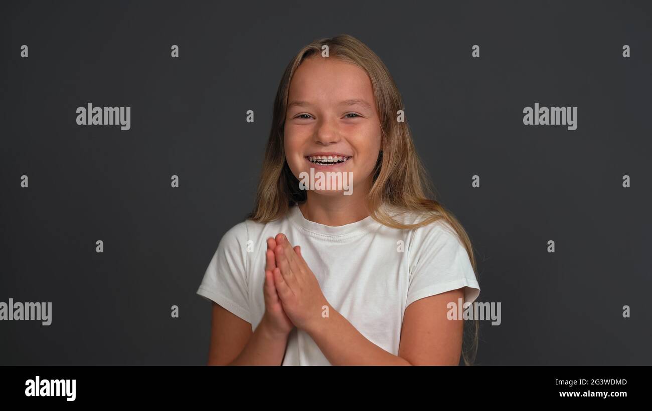 Happy charming little girl with hands put together looking a side of camera, wearing white t-shirt isolated on dark grey or blac Stock Photo