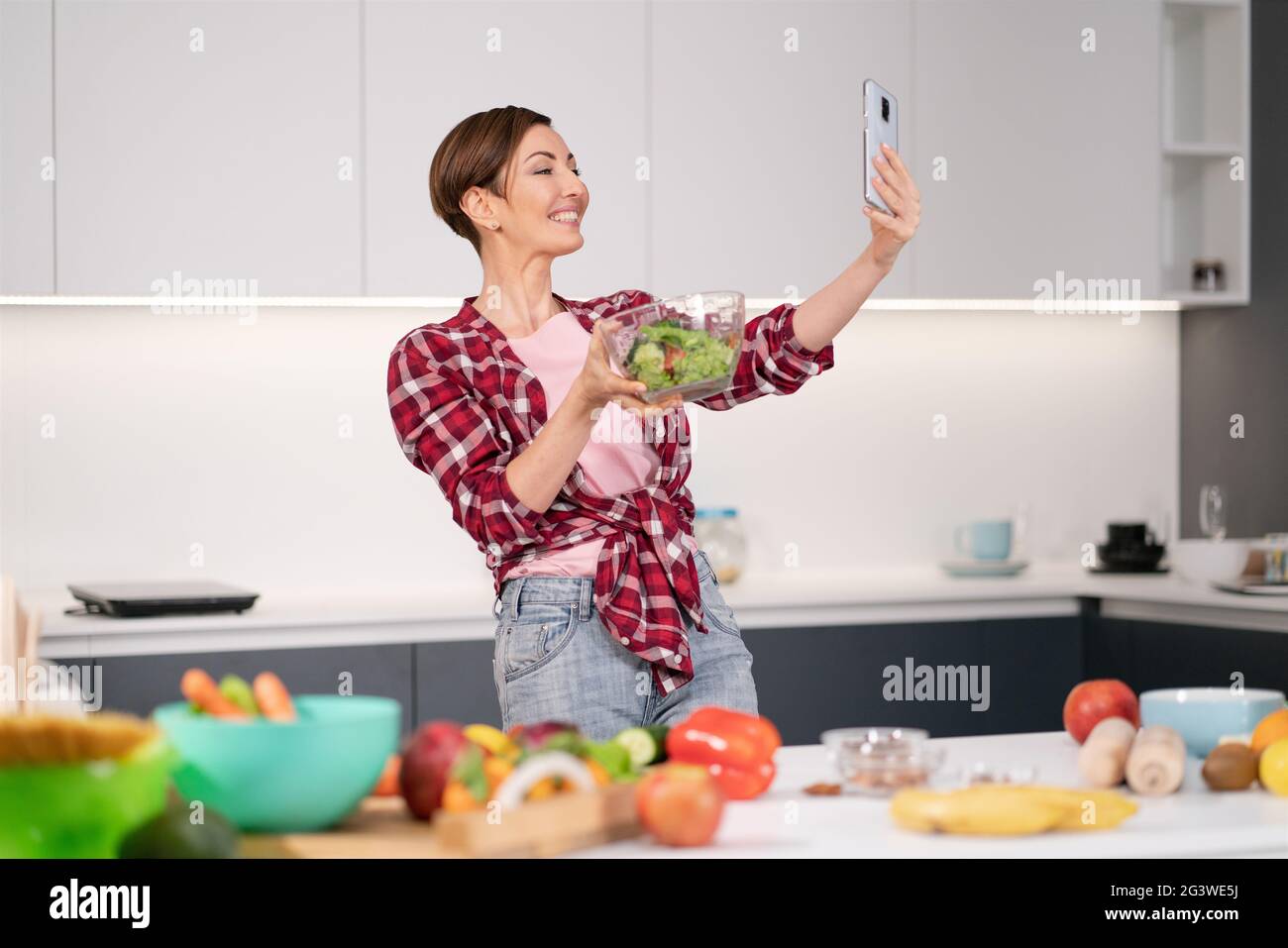 Pretty woman happy taking selfie using her smartphone while cooking fresh salad wearing a plaid shirt with a bob hair style. Hea Stock Photo