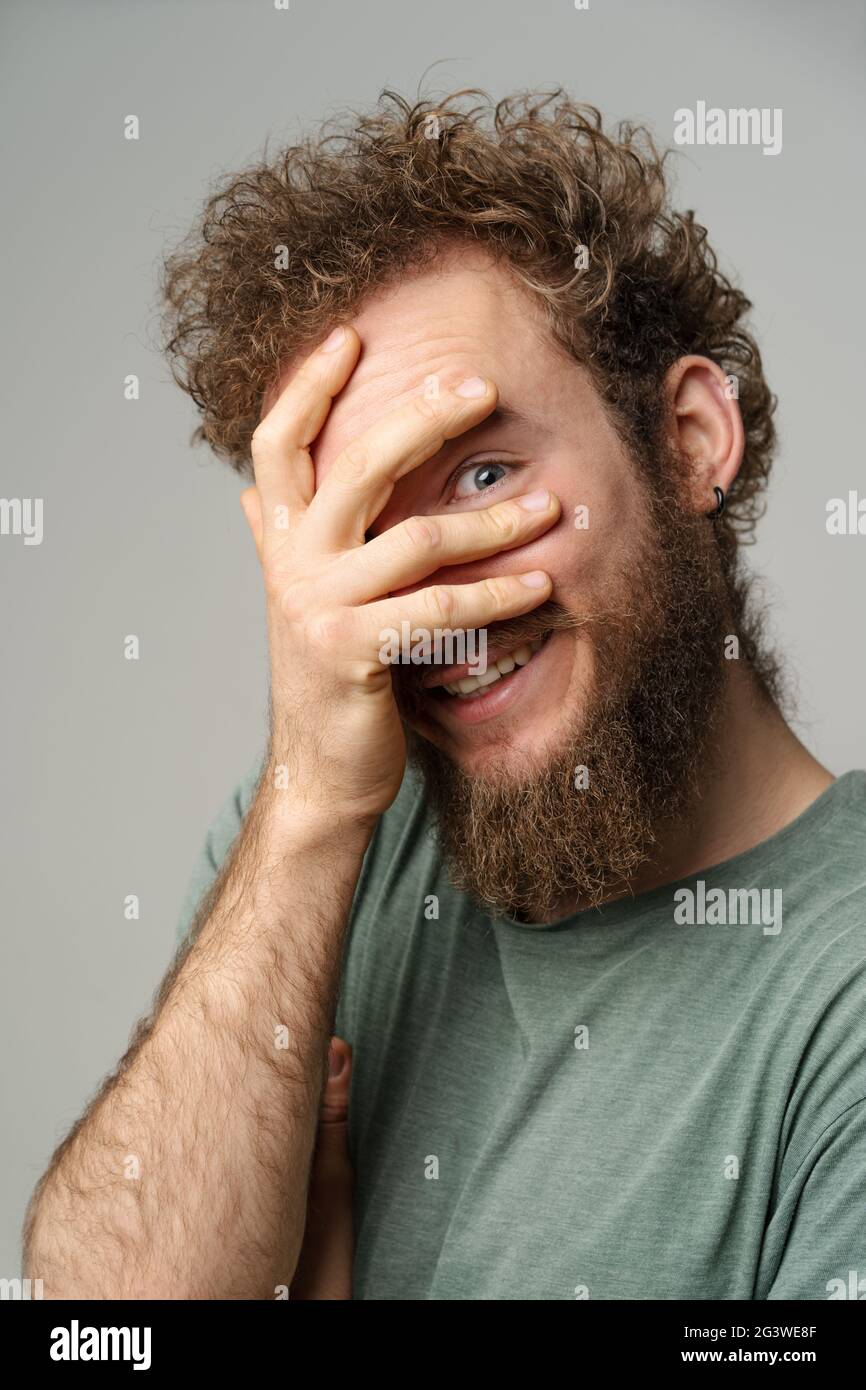 Shy handsome young man covered his face with hand pry with his eye, curly hair in olive t-shirt isolated on white background. Po Stock Photo