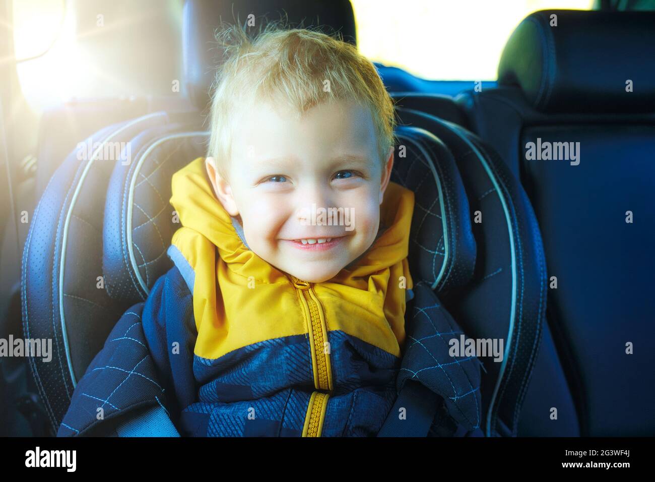 A blond toddler sits in a child's car seat and smiles. This is the happy boy in the car. Care and safety while driving. Stock Photo