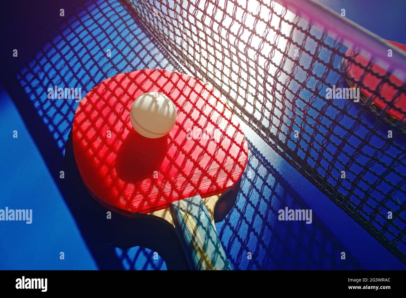 A red table tennis racket and a white ball lie on the surface of the table next to the net. Sports game Stock Photo