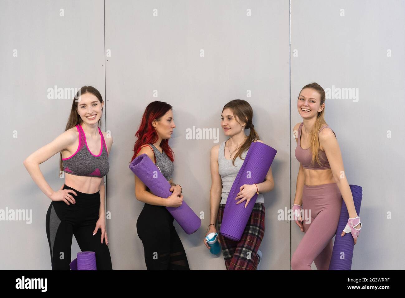 Athletic girls in sports out fits standing next to the wall holding a yoga mat, smiling on camera. Practicing in fitness or yoga Stock Photo