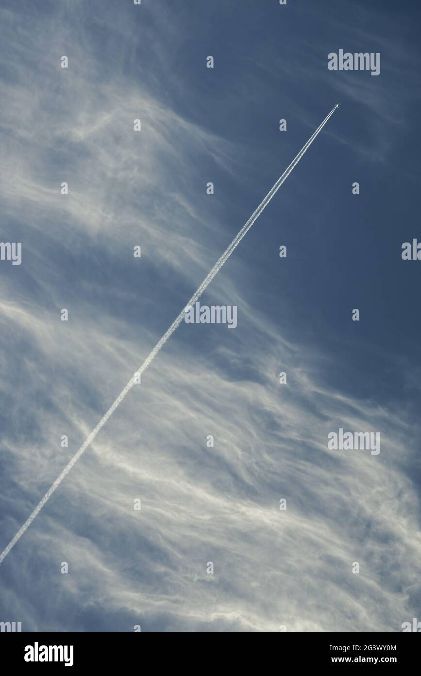 An airplane pits through a blue sky dotted with filamentous white cirrus clouds leaving a brilliant trail Stock Photo