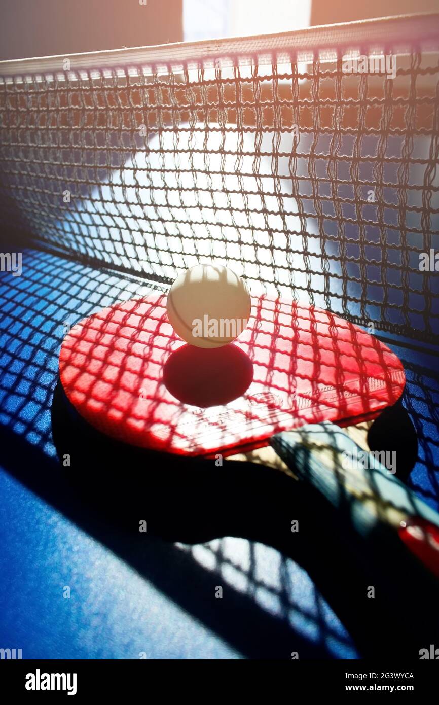 A red table tennis racket and a white ball lie on the surface of the table next to the net. Sports game Stock Photo