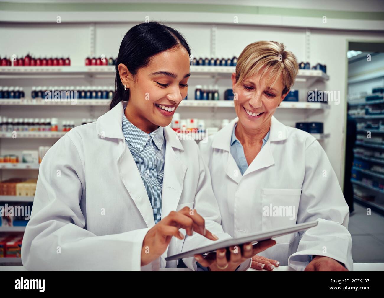 Young woman using a digital tablet and showing something to her senior colleague wearing labcoat while working in pharmacy Stock Photo