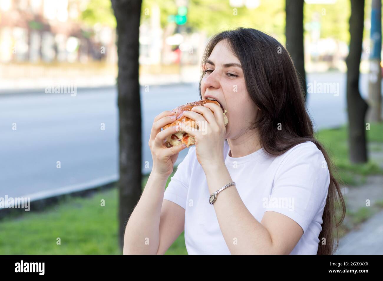 Brunette woman overeats on a burger on the street. Gluttony, excess calories, and bulimia. Stock Photo