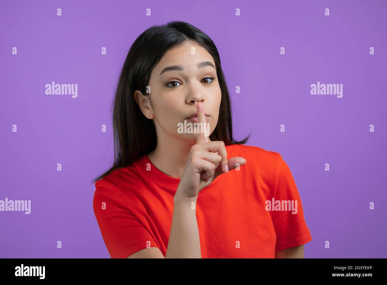 Smiling woman holding finger on her lips over violet background. Gesture of shhh, secret, silence. Close up. Stock Photo