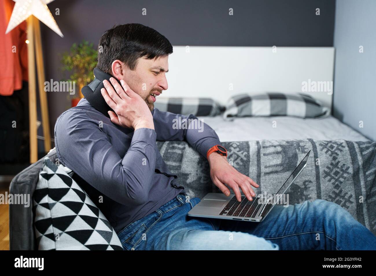 Tired strained man suffering from neck pain while working at computer. Male touchingly massages neck, puts on neck collar, sitti Stock Photo