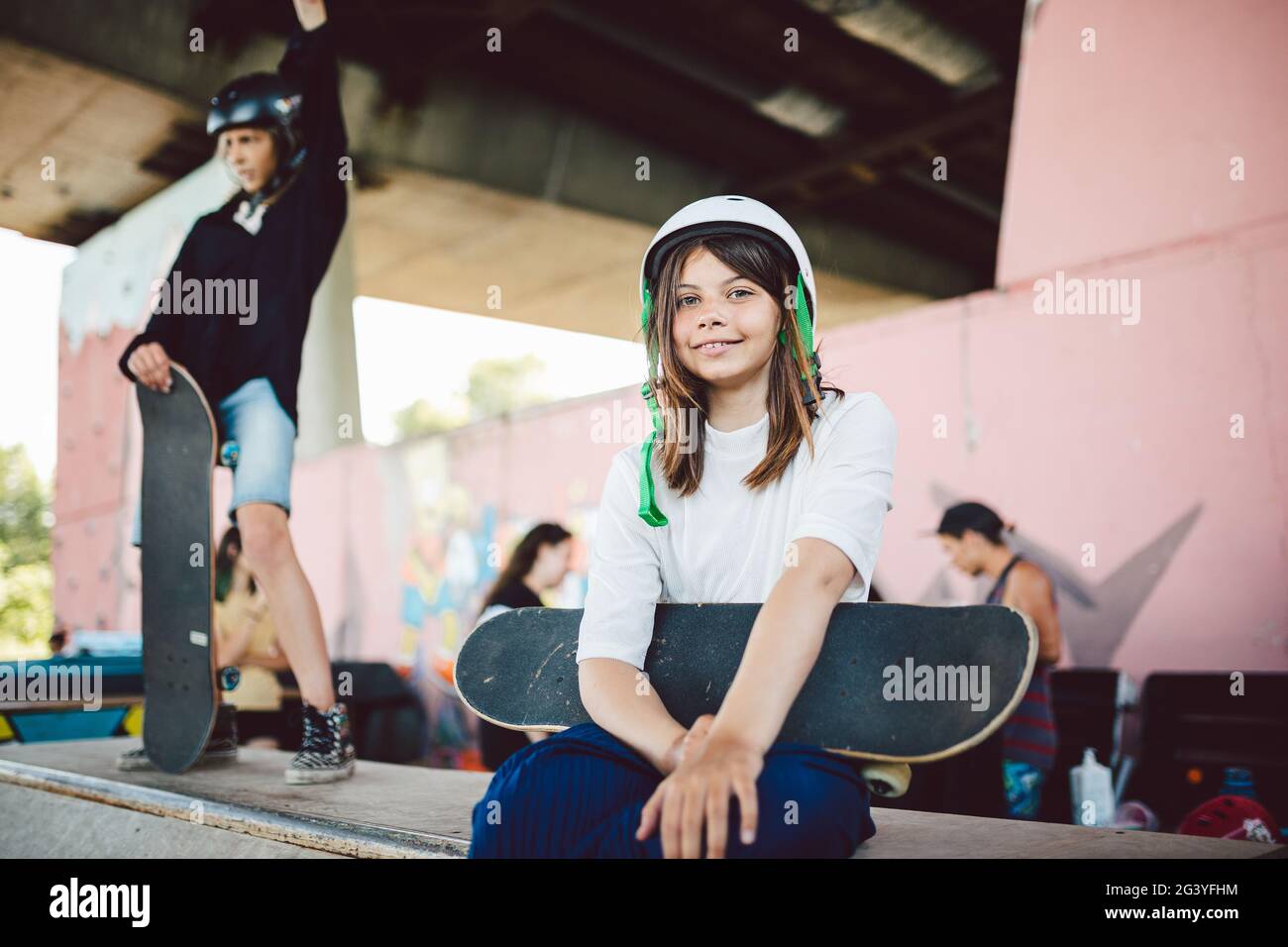 Beautiful teenage girl in white helmet sits with skateboard in her hands on pipe ramp in an extreme sports park in city during t Stock Photo