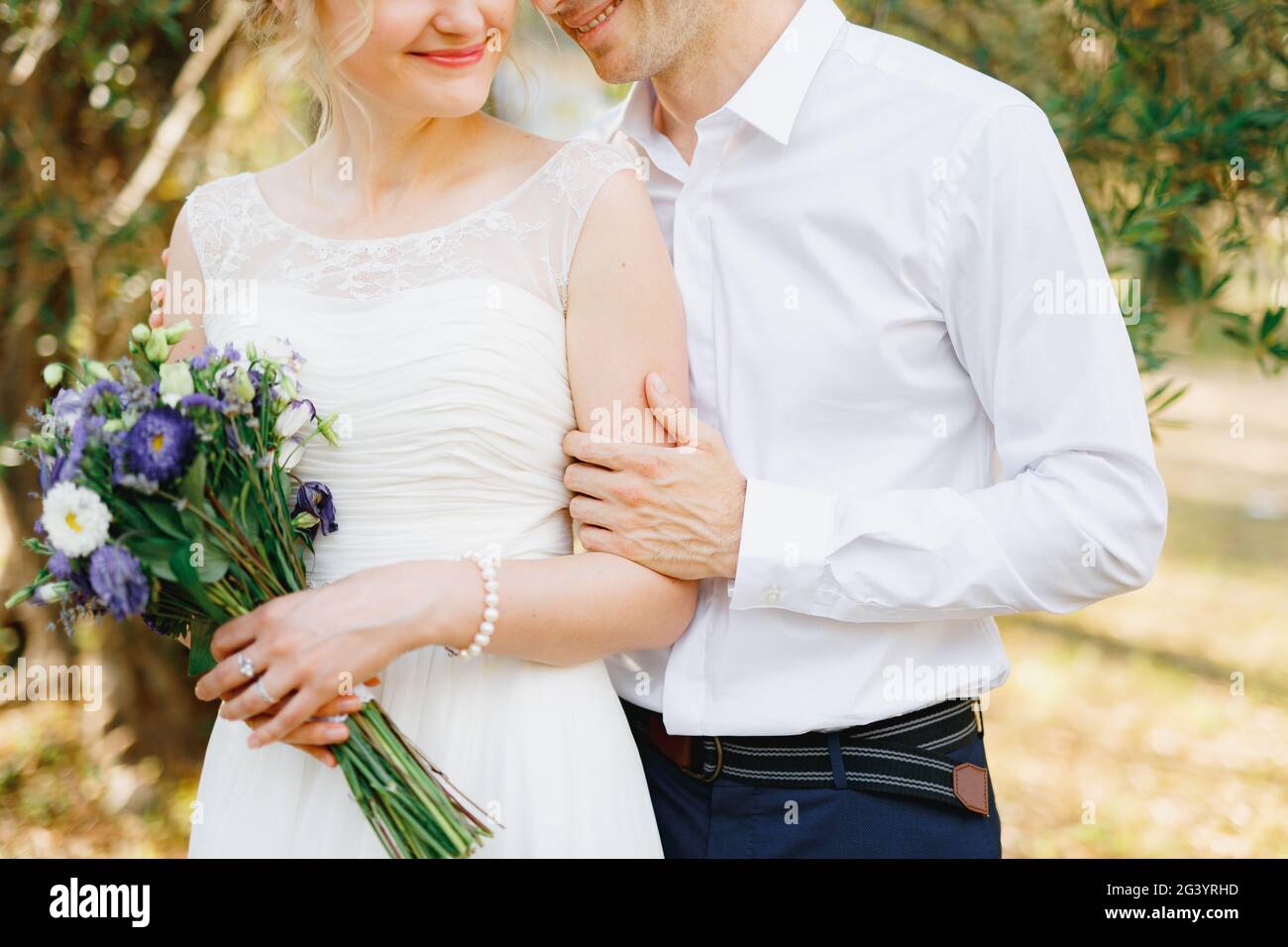 The groom gently hugs the bride near the green trees in the olive grove ...