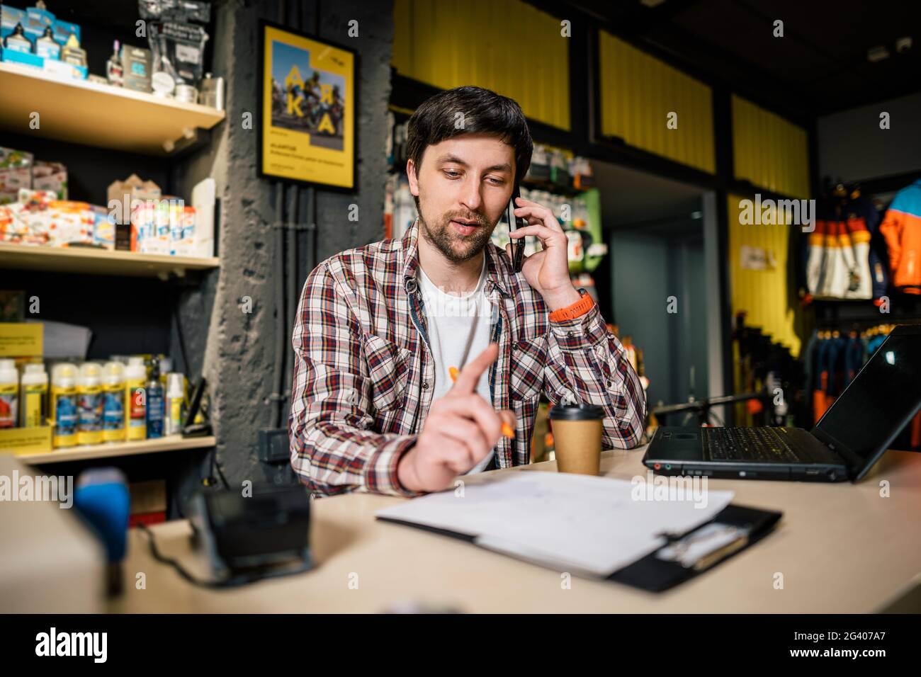 Bike shop owner at work. Store employee takes an order by mobile phone at table near laptop in travel and sporting goods store. Stock Photo