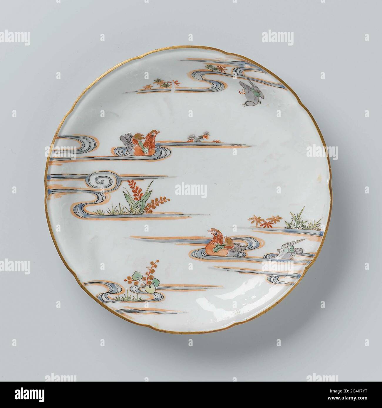 Saucer-dish with Flower Sprays in Relief and Two Mandarin Ducks in a pound. Scale of porcelain with a scalloped edge, painted on the glaze in blue, red, green, yellow, eggplant, black and gold. On the front on the wall flower branches in relief; Two mandarin people in a pond with plants and a flying bird in an email colors. The outside with flowering aquatic plants. Five preen on the bottom. Kakiemon style. Stock Photo