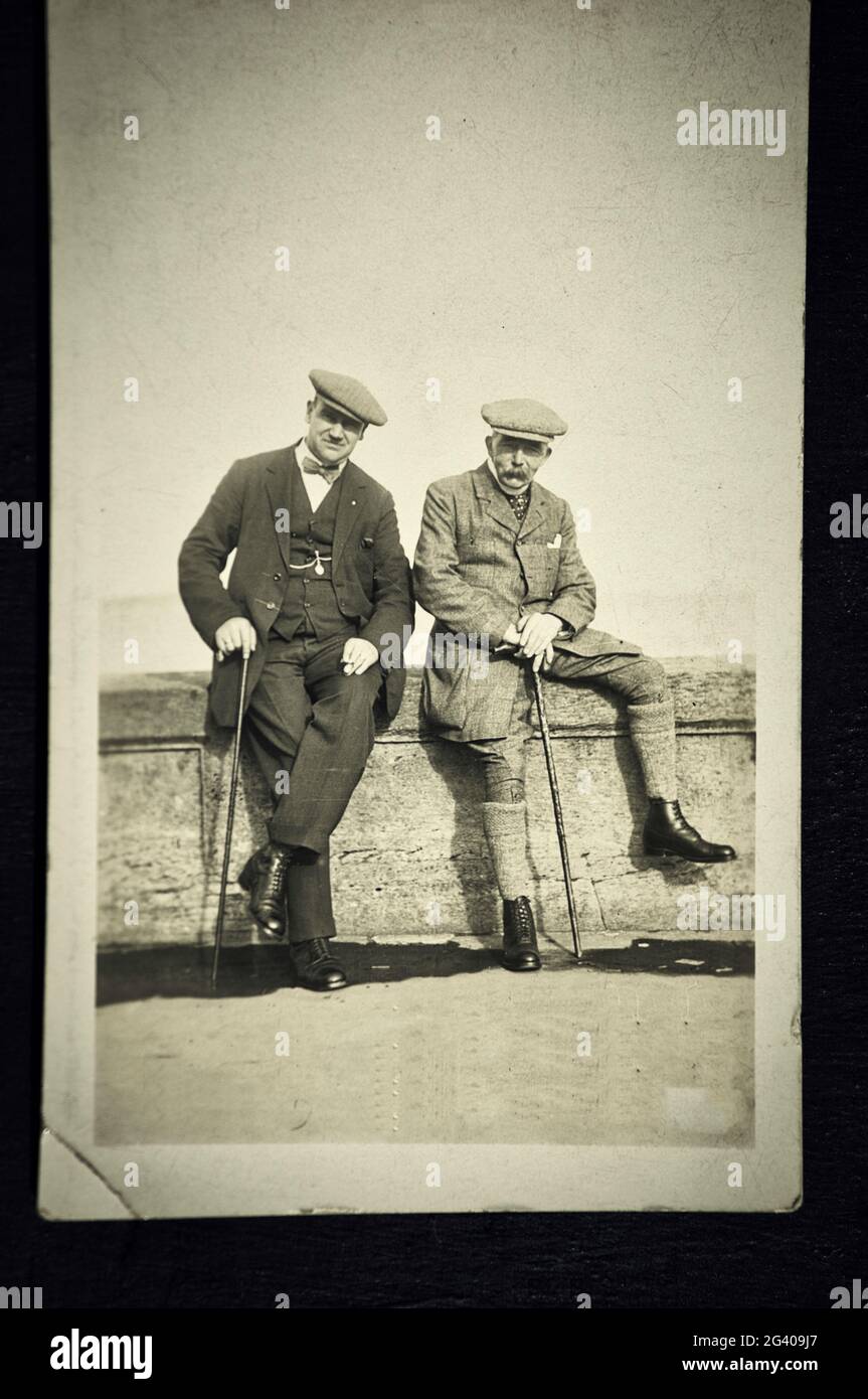 Authentic vintage photograph of two senior men in flat caps and holding walking sticks sitting on wall. Concept of nostalgia, friendship, historical Stock Photo