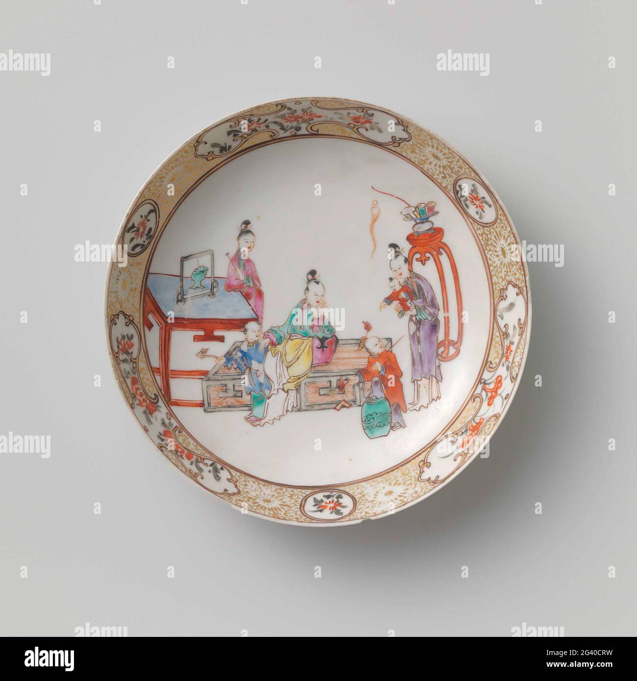 Saucer-Dish With Three Ladies And Children With Precious Objects. Scale of porcelain with round wall, painted on the glaze in blue, red, pink, green, yellow, purple, black and gold. On the front three ladies with three children with valuables; A lady standing next to a table, a lady sitting with a child with scepter, a child with a music stone climbs on a stool, a lady with a child on the arm, behind her a high stool with vase; The inner edge with flower drinks interspersed with round and erratic shaped cartouches with flower branches. A chip and a crack in the edge. Family Rose. Stock Photo