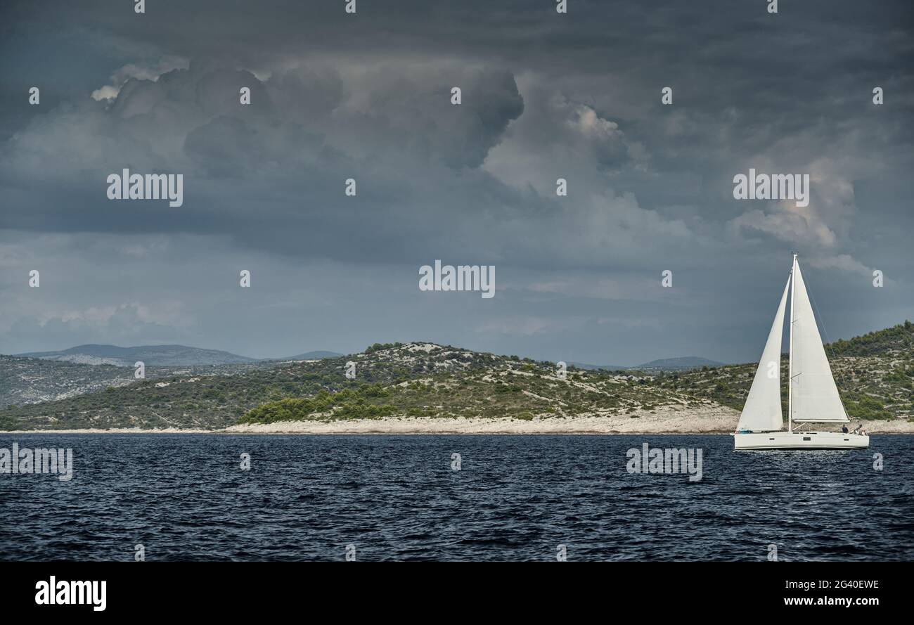 Sailboat of white color, reflection of sail is on water, clouds, the island is on background, sunny weather Stock Photo
