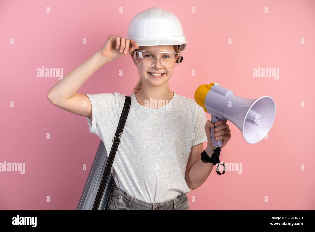 Young engineer is a girl on a background of a pink wall, she is wearing a helmet, glasses and a loudspeaker in her hands. Construction concept. Stock Photo